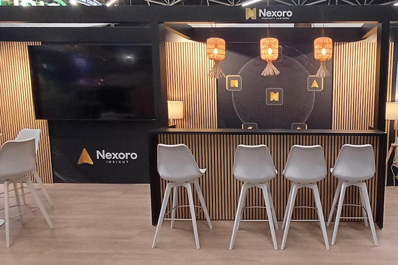 Nexoro's modulaire beursstand met systeemkaders en balie, beide afgewerkt met onze modulaire invulpanelen met houten latjes. De vloer is loose lay. Dat lijkt op vinyl, maar het zijn pvc stroken die los gelegd worden en daardoor herbruikbaar zijn.